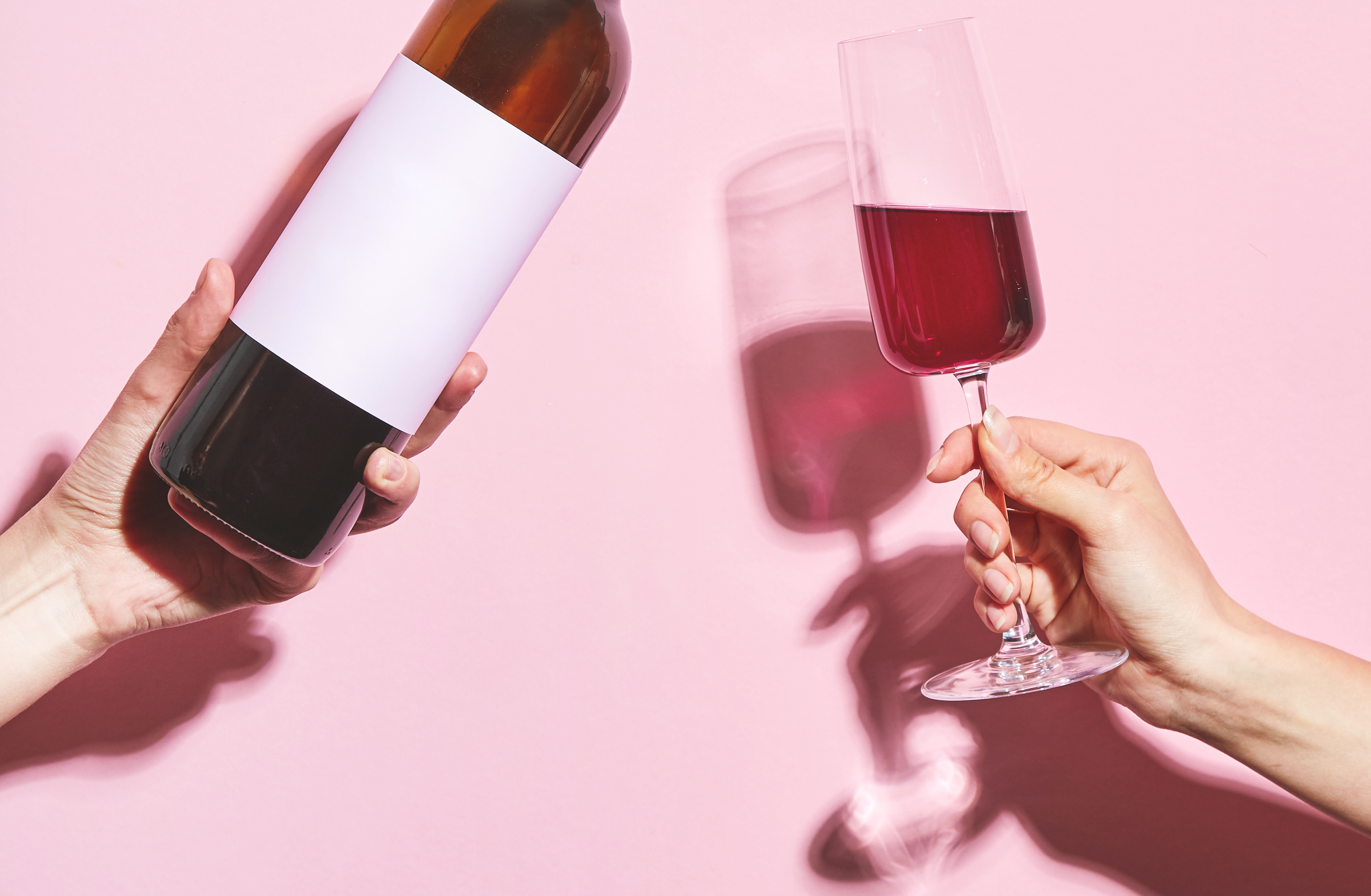 Hands holding a glass of wine and a bottle on pink background. Glass of wine in female hand. Party insta time.