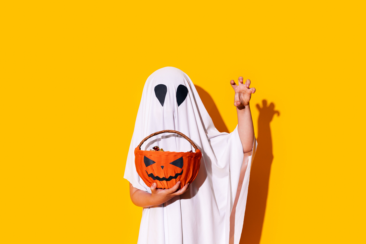 Child in ghost costume holding basket of chocolates in one hand, and raised the other hand up